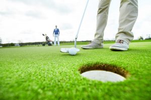 Active man standing on green lawn while holding golf club close to ball before hitting it into hole